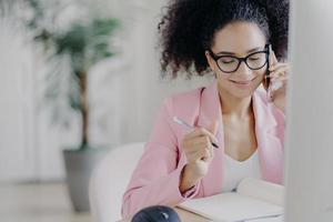 mujer encantadora sonriente con cabello rizado, escribe información en un cuaderno, recibe asesoría por teléfono celular, usa anteojos y traje elegante, trabaja ocupada, es propietaria de una empresa comercial, resuelve problemas foto