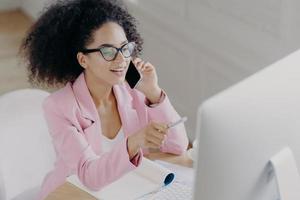 una joven afroamericana alegre habla de noticias de negocios, señala con un bolígrafo en la pantalla de la computadora, habla a través de un teléfono celular moderno, está ocupada con el papeleo, usa anteojos transparentes y ropa formal foto