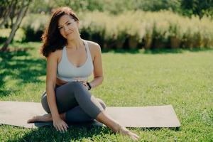 Photo of brunette sportswoman has rest after fitness training sits on karemat with bare feet in activewear leads healthy lifestyle poses on fresh green grass enjoys summy day and good weather