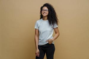 Positive ethnic female student feels free after passing exam, smiles broadly, keeps hand in pocket of jeans, wears transparent glasses, poses against brown background. People, lifestyle concept photo