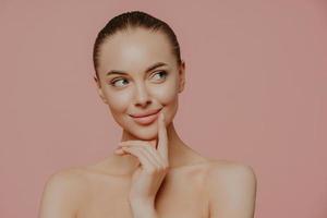 Topless thoughtful lady with clean perfect skin, dark hair, looks aside, undergoes facial trearrments, poses indoor, isolated over rosy background. Cosmetology, skin care and wellness concept photo