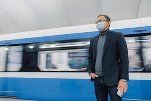 Pensive businessman wears surgical sterile mask to prevent spread of respiratory disease, poses against underground train, travels in public transport, holds smartphone in hand. Coronavirus outbreak. photo