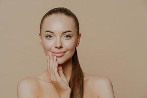 Close up portrait of beautiful naked young woman smiles tenderly and touches face, has healthy clean skin, cares about appearance, poses against beige background. Youth and skin care concept. photo