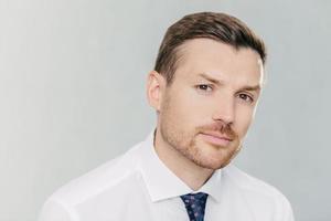 Close up portrait of handsome unshaven man looks seriously at camera, has appealing look, thinks about business affairs, dressed in formal clothes, isolated over white background. CEO indoor photo