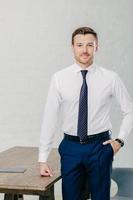Photo of handsome male office worker dressed in formal clothes, keeps one hand in pocket, other on table, looks confidently at camera, poses in office. People, business and lifestyle concept
