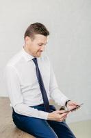 Young unshaven intelligent male wears formal clothes, uses digital tablet for work, focused into screen, sits at office desk, isolated over white background. Programmer develops application software photo