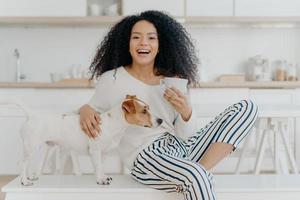 People, leisure and animals concept. Joyful dark skinned woman petts favourite dog, drinks hot beverage from white cup, looks happily aside spend time in kitchen. Caring host with jack russell terrier photo