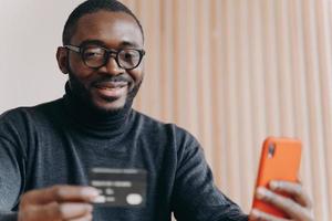 Young positive African ethnicity man entrepreneur in glasses paying with credit card online photo