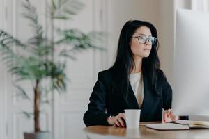 Concentrated female entrepreneur wears elegant black costume, works at computer, sits at table in cozy work place wears transparent glasses browses internet makes business project thinks over strategy photo