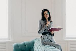 Serious businesswoman dressed in formal wear, concentrated into distance, ponders on creative ideas, writes down notes in notepad, wears spectacles, poses on back of couch in spacious light room photo