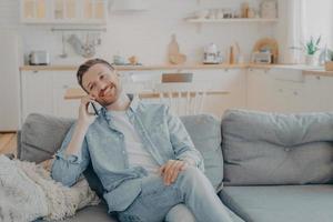 Smiling guy sits on couch at home having fun while talking on modern cellphone gadget photo