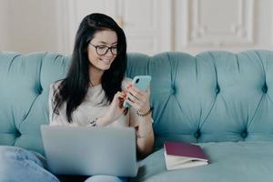 Successful brunette female freelancer watches live stream video online, poses at comfortable sofa with modern gadgets, reads message on cellphone, works distantly from home, dressed casually photo