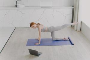 Young happy european woman is doing plank exercise and looking into laptop. Workout online at home. photo