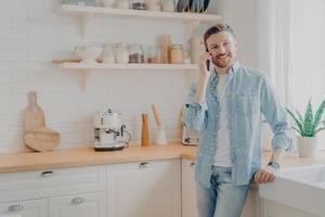 apuesto joven barbudo sonriente con ropa informal hablando por teléfono móvil foto