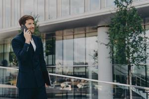 toma lateral de llamadas de hombre barbudo a través de un teléfono inteligente vestido con traje formal negro posa al aire libre
