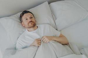 Lonely upset young bearded man lying in bed with open eyes photo