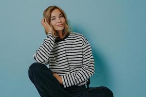 Photo of attractive young woman with minimal makeup sits relaxed on chair over blue background