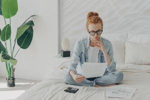 mujer europea de jengibre enfocada sentada en la cama y calculando facturas domésticas en casa, sosteniendo papeles foto