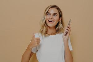 Young satisfied girl in casual clothes holding mobile phone and showing thumb up gesture, being excited photo