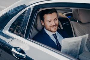 Handsome young banker in smart formal tuxedo suit reads newspaper in luxury auto