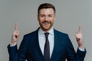 Pleased businessman in suit pointing up with fingers at copy space photo