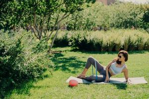 Outdoor shot of slim sporty young woman in activewear practices exercises for legs with elastic band lying on karemat at green grass enjoys fresh air keeps fit. People fitness aerobics concept