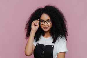 Pretty African American woman keeps hand on frame of glasses, smiles happily, wears white casual t shirt and black overalls, enjoys spare time, poses over violet background. Ethnicity, fashion concept photo