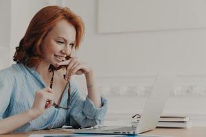 Pleased female model poses at coworking space, holds spectacles, smiles positively, uses wireless internet connection, reads information, has online communication, works on creating web project photo