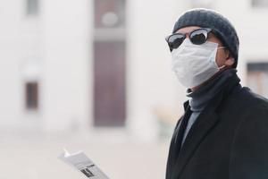 Horizontal shot of man being on quarantine, wears sunglasses and protective medical mask, walks outdoor, cares about health during global coronavirus epidemic, reads press to know fresh news photo