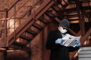 Coronavirus precautions. Male model avoids crowded places, fights against new virus, reads attentively article about covid-19 symptoms, holds newspaper, wears protective face mask and medical gloves