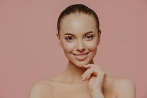 Studio shot of pretty charming woman touches chin, has healthy glowing skin, cares about complexion, stands naked indoor, has cosmetology procedure, isolated on pink background. Beauty concept photo