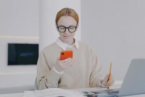 Happy young woman is texting at workplace in office. Girl is sending messages at her desk. photo