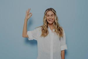 Young joyful blonde caucasian woman demonstrating agreement and showing okay gesture with closed eyes photo