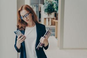 Redhead business woman with serious look in eyewear using cell phone while standing in office photo