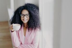 Indoor shot of cheerful curly haired African American lady has coffee break, holds white cup of drink, wears optical spectacles, formal outfit, stands near white wall, works in business sphere