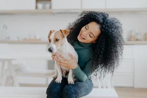 feliz propietaria del perro jack russell terrier, siente la responsabilidad de cuidar a la mascota, tiene el pelo rizado oscuro y tupido, se sienta contra el fondo borroso de la cocina. las personas y la relacion con los animales foto