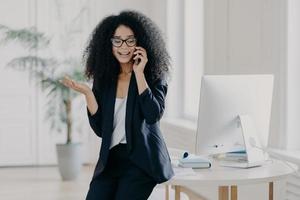 Photo of puzzled prosperous businesswoman calls partner, raises palm, holds mobile phone, wears spectacles and formal black outfit, tries to solve problem at work, discusses something actively