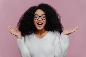 Portrait of joyful curly lady raises arms and spreads palms, wears optical glasses and white sweater, gets pleasant unexpected surprise from close person, isolated on violet studio wall. Happiness photo