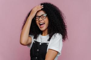 Una mujer de pelo rizado, muy contenta y complacida, mantiene la mano en la frente, se ríe de la felicidad, entiende que hizo algo divertido, usa anteojos ópticos, ropa informal, modelos con fondo morado foto