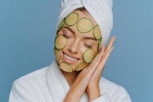 Relaxed satisfied woman tilts head on palms pressed together keeps eyes shut and smiles gently makes facial mask applies cucumber slices on face enjoys morning skin care routine poses indoor photo