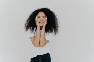 Candid shot of good looking lady with curly hairstyle, smiles cute, keeps both hands on cheeks, shows positive emotions, wears white t shirt, black denim trousers, stands indoor, blank copy space photo
