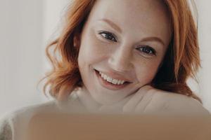 Close up shot of positive redhead woman with freckled skin and toothy smile, concentrated at display of laptop computer, satisfied with online business, checks received message, has remote job photo