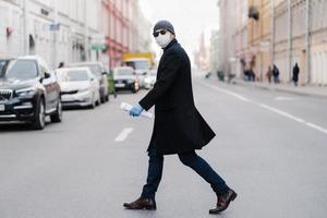 Virus covid -19. Man crosses road, dressed in black coat, hat and sunglasses, wears ptotective mask for pandemic virus protection, poses outdoor at busy street with many transport and people photo