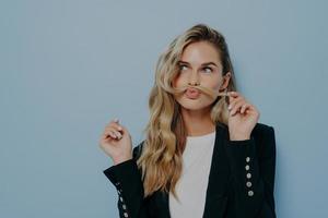 Funny joyful blonde woman having fun while posing against blue studio wall, playing with strand of hair photo