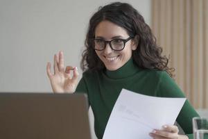 Smiling Italian female employee holding financial report and showing okay gesture during video call photo