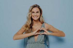 Happy lovely female with blonde long hair showing love symbol with her hands cupped in heart shape photo