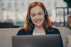 una joven pelirroja sonriente y complacida con auriculares hablando con colegas en línea en una laptop foto