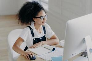 una joven afroamericana contenta se sienta frente a una computadora moderna, hace que el proyecto funcione, tiene muchos papeles y un bloc de notas en la mesa, usa anteojos para corregir la visión, se viste con ropa informal foto