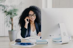 foto de una mujer afroamericana satisfecha llama a través de un teléfono inteligente, toma notas en el bloc de notas, participa en el proceso de trabajo, se sienta en el escritorio con la computadora, usa anteojos, ropa formal, tiene comunicación telefónica