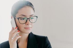 una foto en la cabeza de una gerente morena seria con maquillaje usa anteojos de moda, ropa formal, tiene manicura roja, llama a un socio de negocios durante un trabajo remoto, posa en el interior con fondo blanco